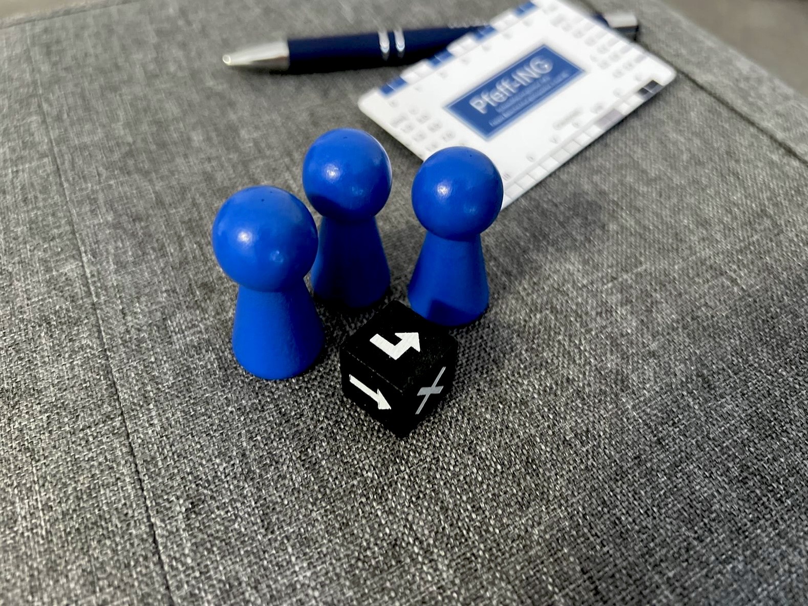 Drei blaue Spielfiguren, ein schwarzer Würfel, ein Kugelschreiber und eine Prüfkarte mit dem Logo von Pfeff-ING auf grauem Stoffhintergrund.